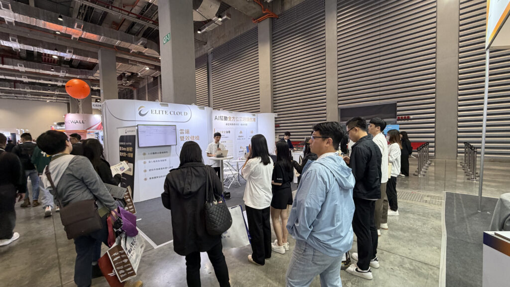 People line up at an exhibition booth labeled 'Elite Cloud' with a presenter and display screens in a large indoor conference hall.",